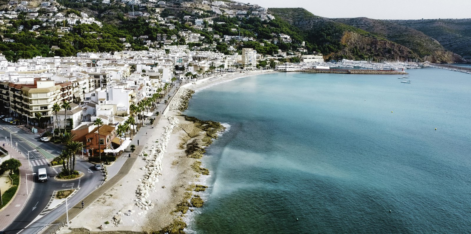 Aerial photo of Jávea's harbour, with the pebble beach of La Grava and the marina, backed by the green flank of Cap de Sant Antoni and the villas of La Corona.